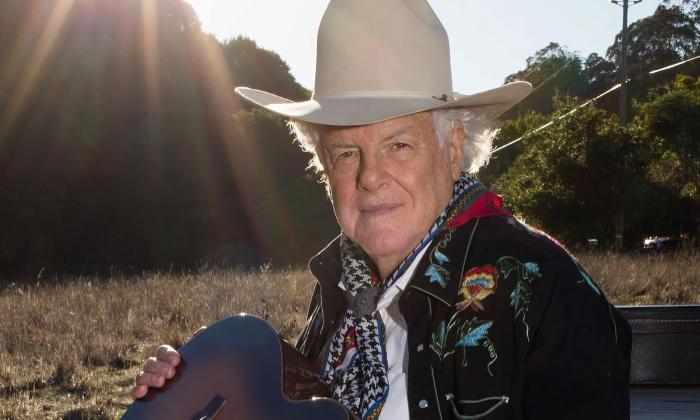 Peter Rowan in a tall white cowboy hat, outside on a prarie