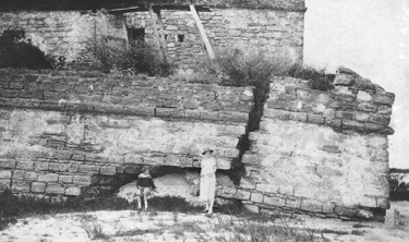 A black and white photograph of Fort Matanzas as it appeared in 1912, in ruins. The gundeck is cracked down the middle and lies crooked, with plants growing atop it. In the foreground, two people pose for the photograph. On the right is a woman wearing a white dress and a hat and to her left is a child, sitting on a large piece of the rubble, in a black garment. It is a small pixelated image.