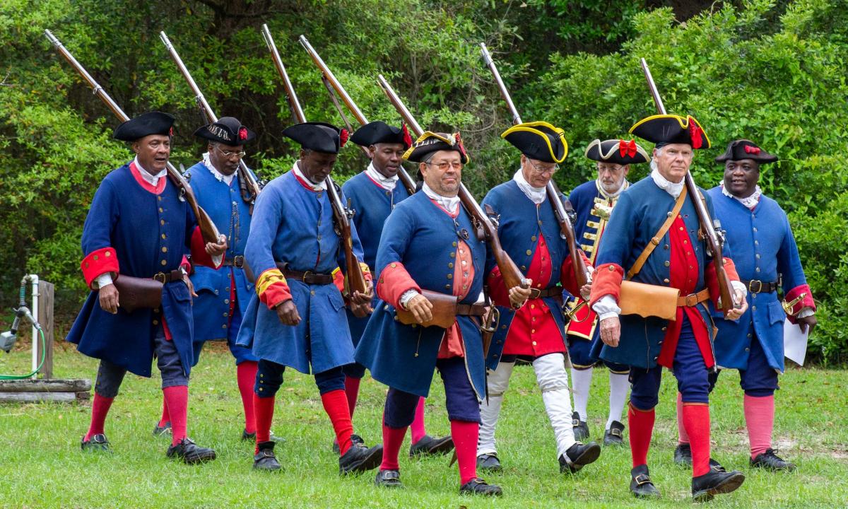 A group of reenactors of different races wearing 1700s Spanish soldier garb march in formation.