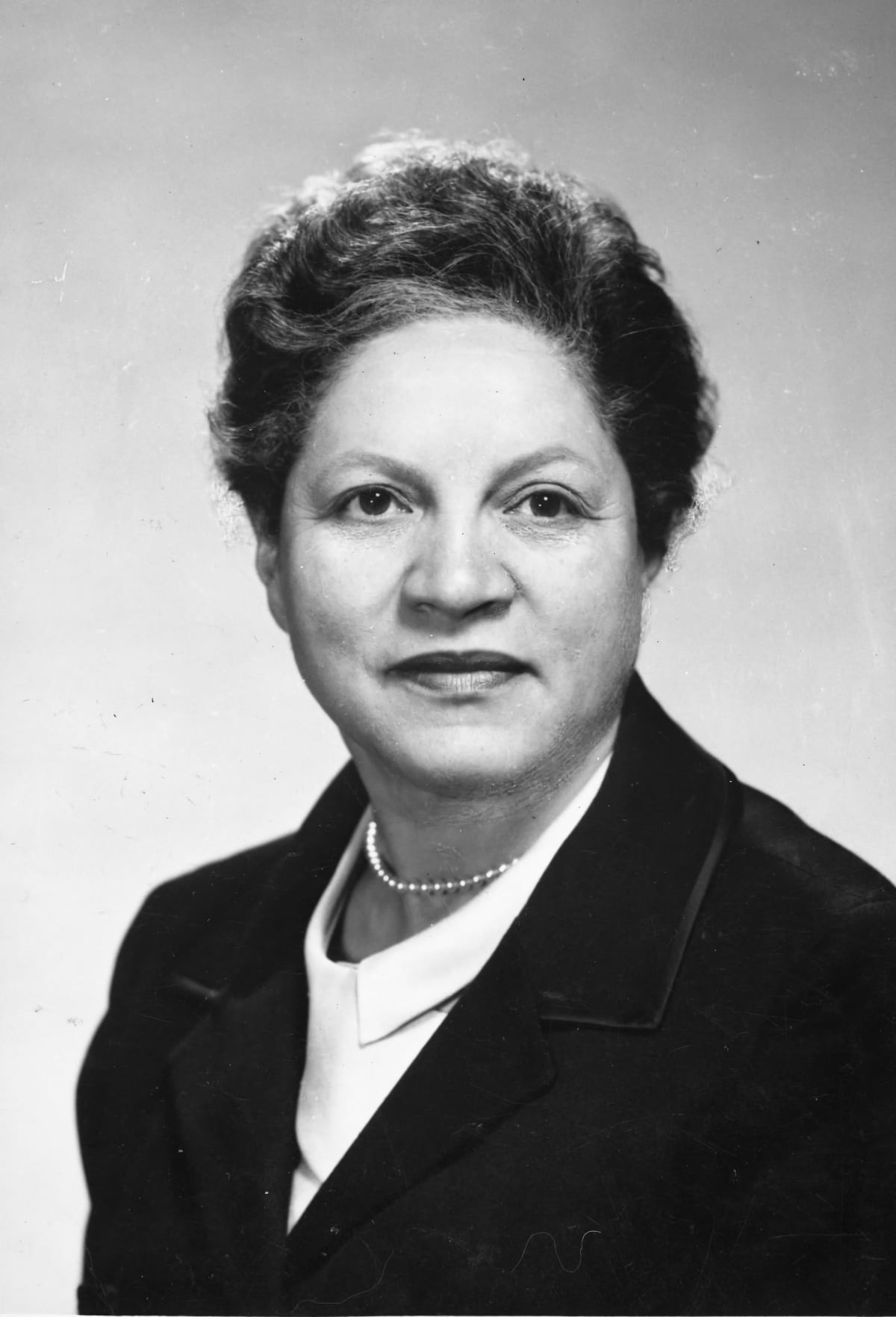 B&W Esther Taylor Burgess portrait. A woman in her 50s in a black blazer wearing pearls.