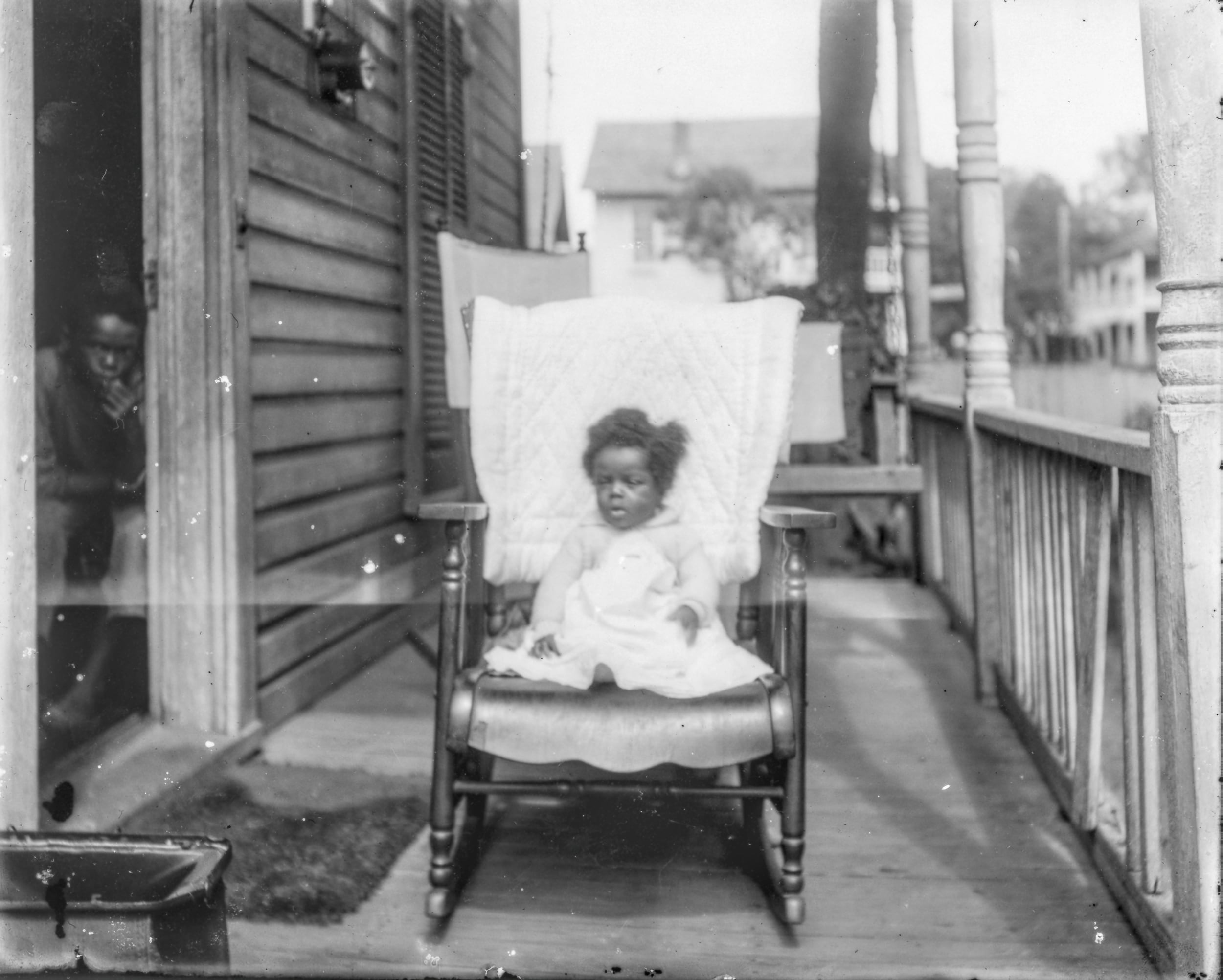 B&W, an African American toddler dressed in white sits on a rocking chair on a Victorian porch. Another child, male, gazes from within the open front door.