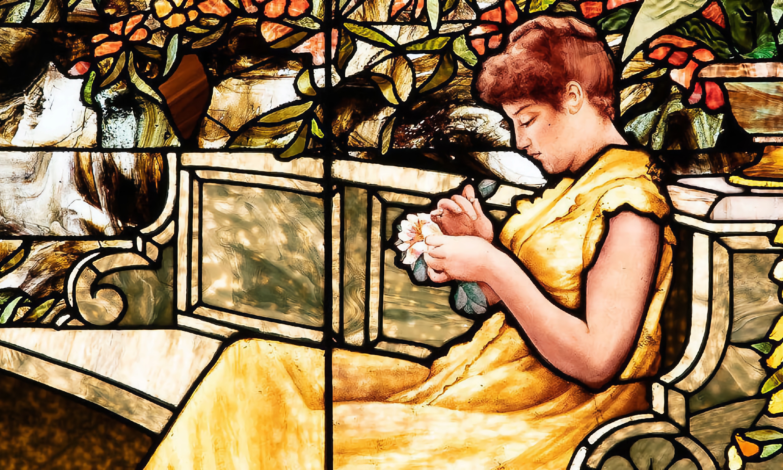 A section of a stained glass window shows a woman in a yellow dress sitting on a garden bench