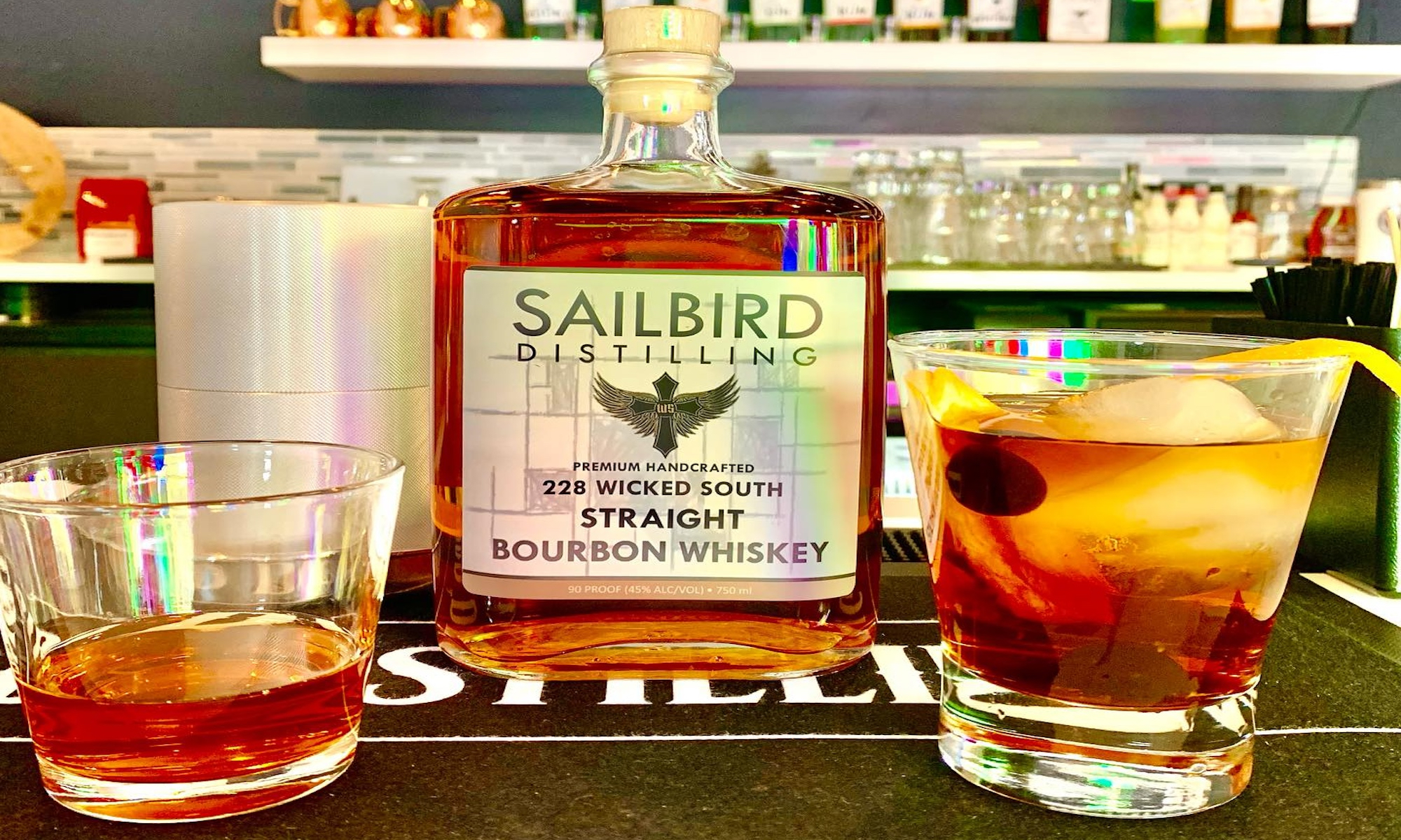 A bottle of bourbon sits on a bar with two bourbon drinks