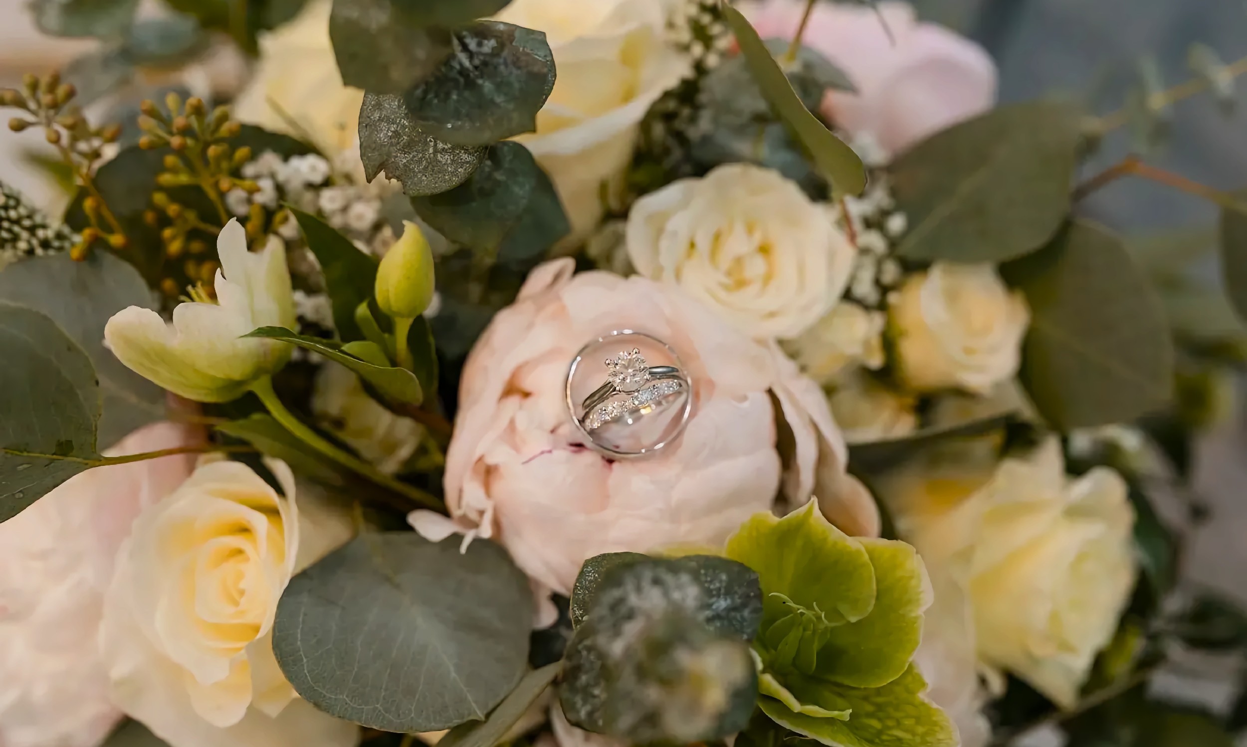 A soft bridal bouquet of pink and yellow roses and greenery, with the couple's rings nestled in a pink rose