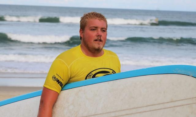 Fitch McGraw, surfing instructor, walking along the beach carrying his board