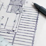 A close up full color image of a drafted drawing of a home's design. A pen and ruler are laid over the paper for aesthetic appeal.