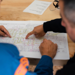 A full color image of two men facing a desk full of construction planning documents. They point to the paper, assumedly making a plan. 