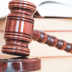 A full color image of a wooden gavel on its stand. Books in the background for academic vibes.