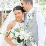 A bridal couple standing outside near water, the bride has a white and green bouquet