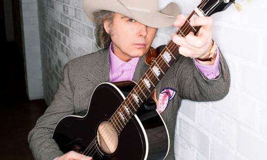 Dwight Yoakam strums his guitar and poses in front of a white wall. 