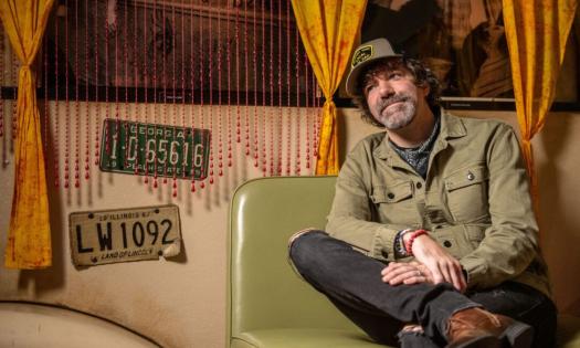 Stephen Kellogg smiles and poses on a green chair with gold curtains as a backdrop. 
