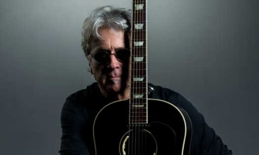 Drew Harrison wears black clothing with matching glasses and clutches his guitar in front of a grey backdrop. 