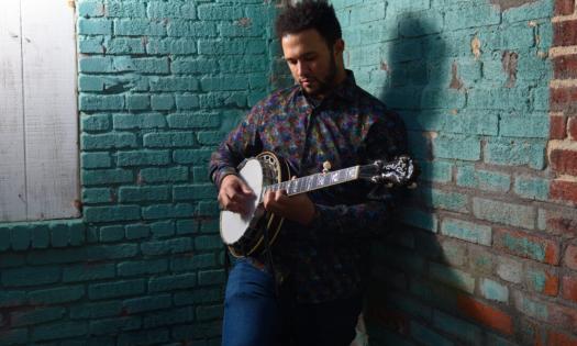 Tray Wellington strums his banjo in front of a brick wall.