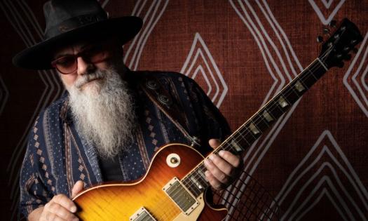 Jimmy Vivino wears a black hat and glasses while posing with his guitar.