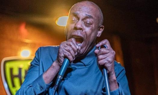 Michael Winslow wears a blue shirt while speaking with a microphone pressed to to his lips.