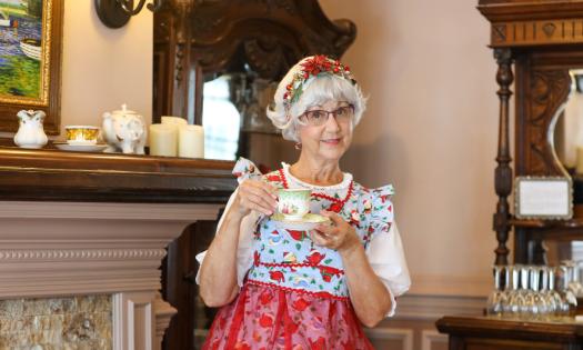 A woman, dressed as Mrs. Sunshine Santa, in a red and blue patterned jumper, holds a coup of tea standing next to a fireplace mantle