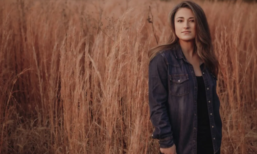 Jennifer Knapp stands against a backdrop of wheat, wearing a black jacket. 