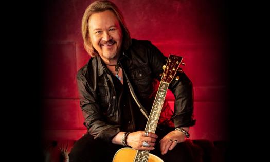 Travis Tritt sits with his guitar against a red and black backdrop.