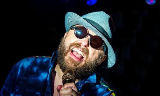 John Nemeth sings while wearing a gray fedora and dark blue shirt.