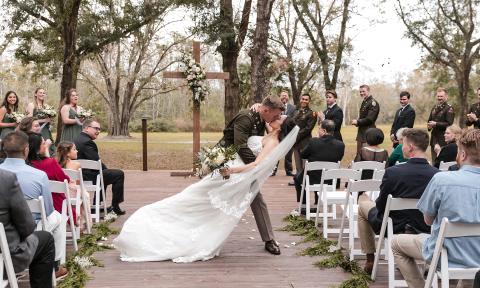 A military groom sweeps his bride into a kiss as the at an outdoor wedding with attendants and guests watching