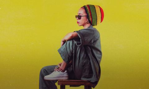 Nattali Rize sits in a chair and poses in front of a bright yellow backdrop. 