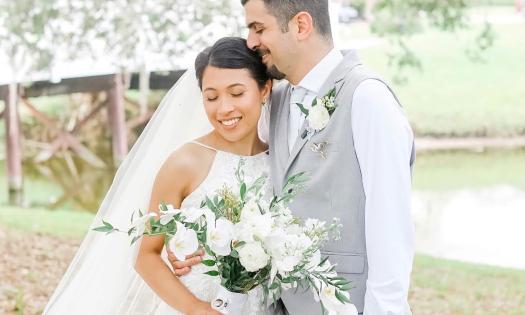 A bridal couple standing outside near water, the bride has a white and green bouquet