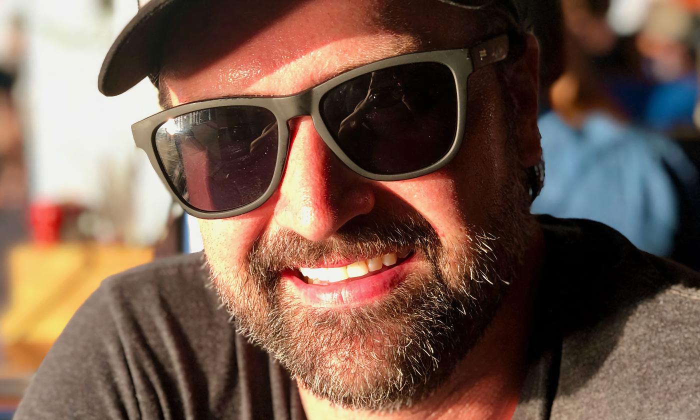 A close-up of a smiling man, wearing sunglasses and sporting a beard and ball cap, outside in a crowd.