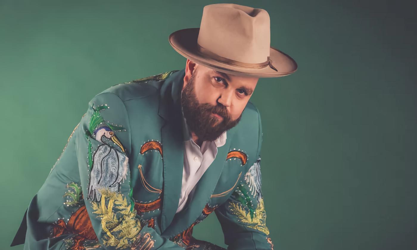 A man wearing an green suit coat, appliquéd with birds, and a brown fedora, against a green background