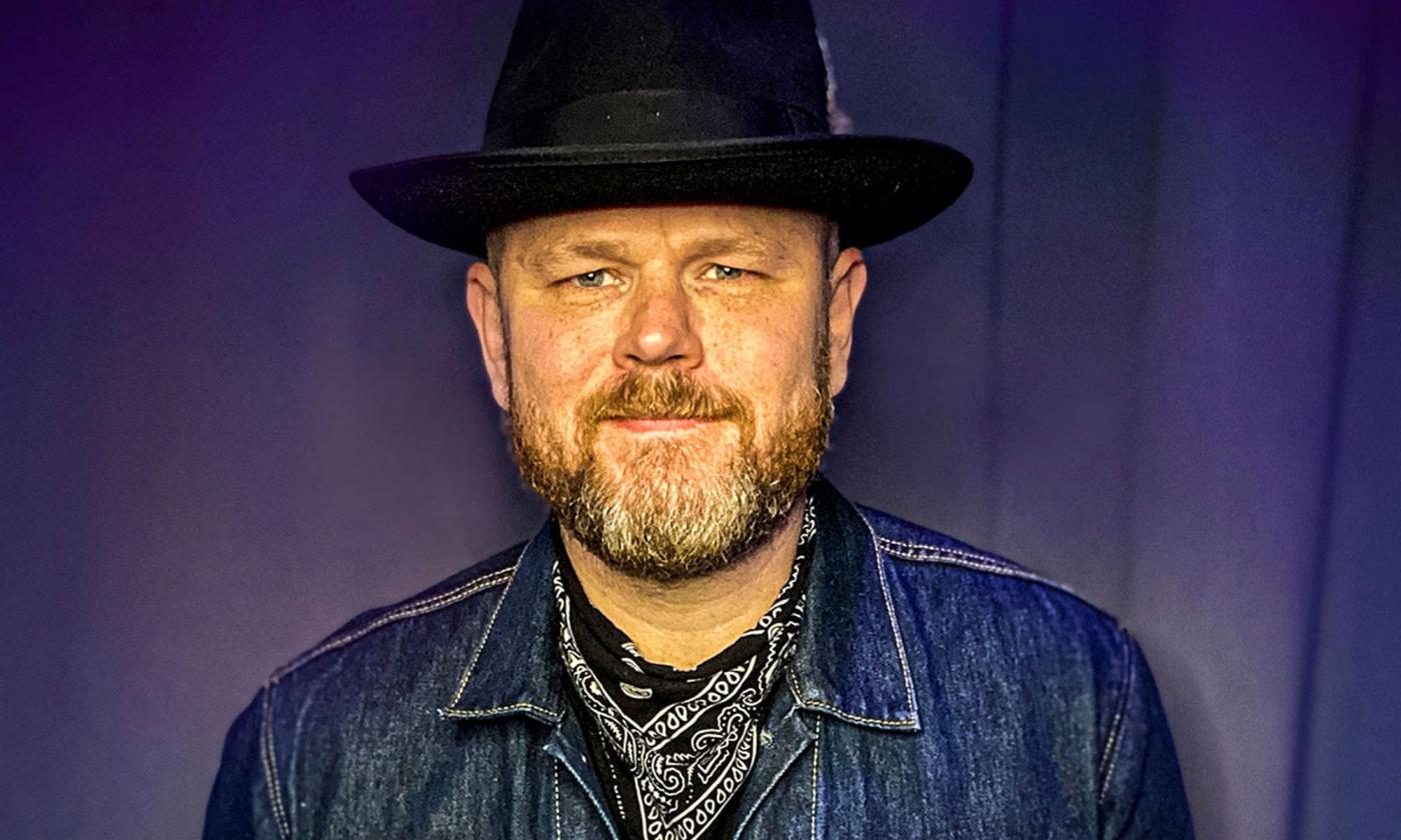 A man with a short beard, wearing an old-fashioned hat and jean jacket, in front of a purple curtain