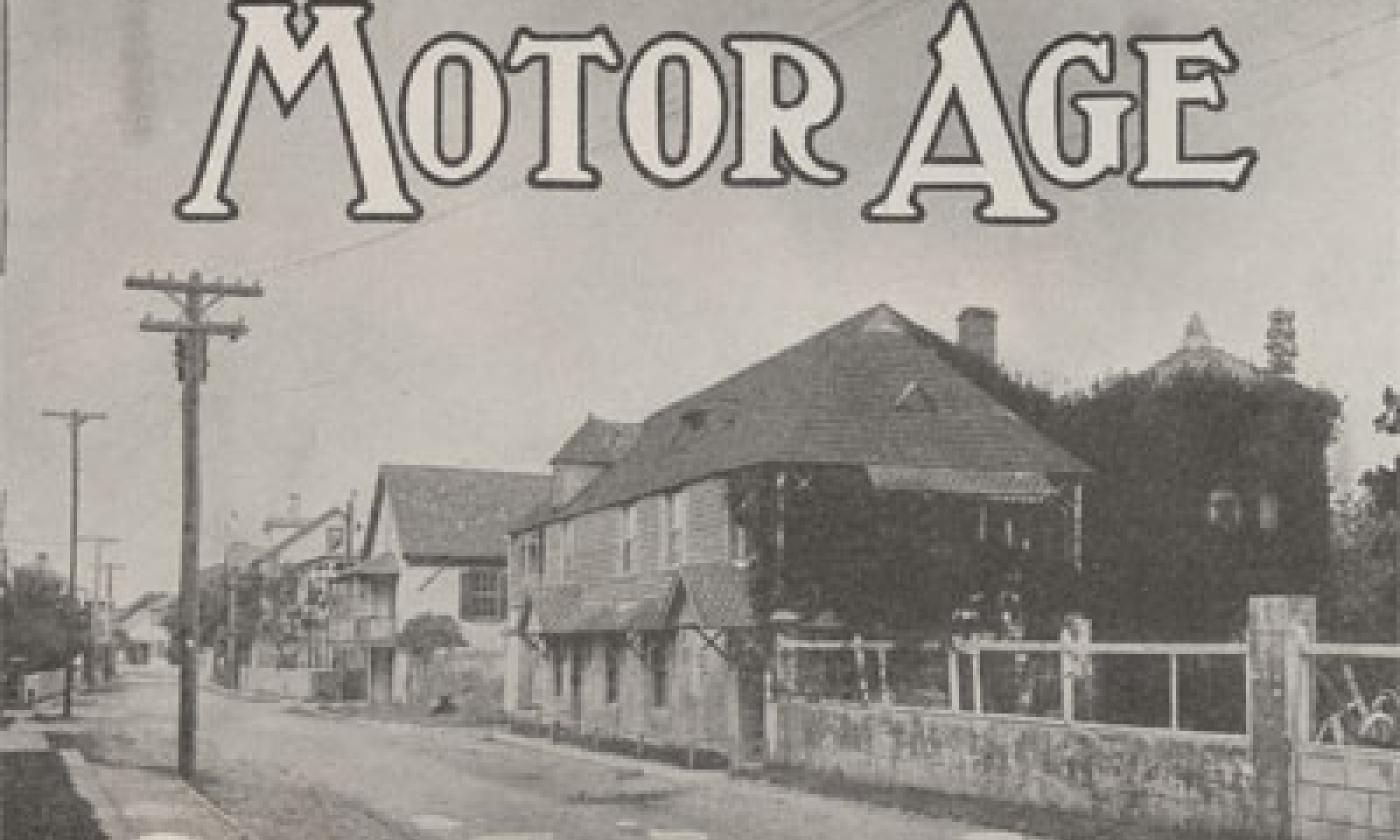 A black and white photograph of a street in St. Augustine, Florida, circa 1915. In the foreground is a coquina wall that runs parallel to the street, which is sand. In the middle of the image is a row of historic houses. Power lines can also be seen. On the top border of the image, the words MOTOR AGE is superimposed over the sky in a serif font.