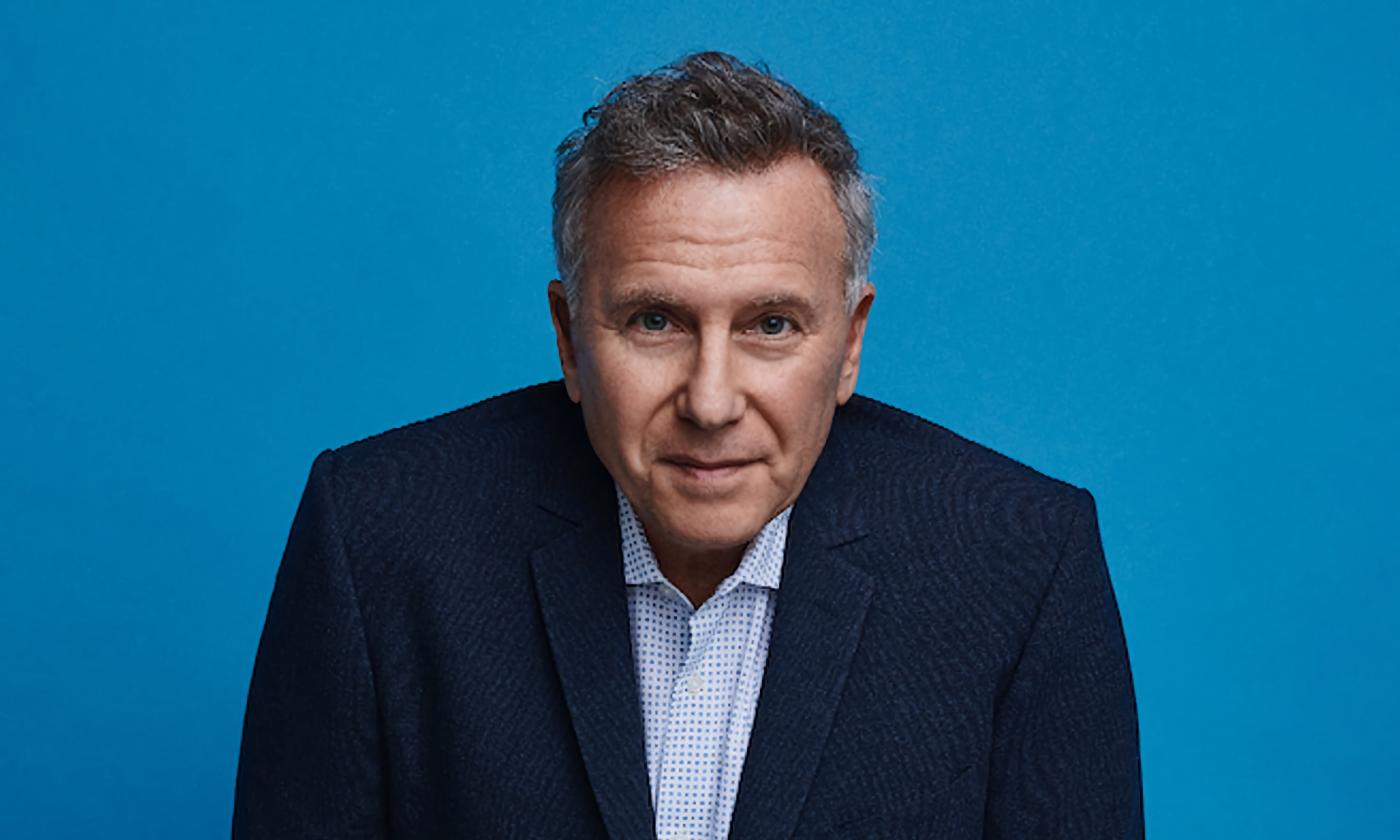 Paul Reiser, in a dark suit jacket against a bright blue background