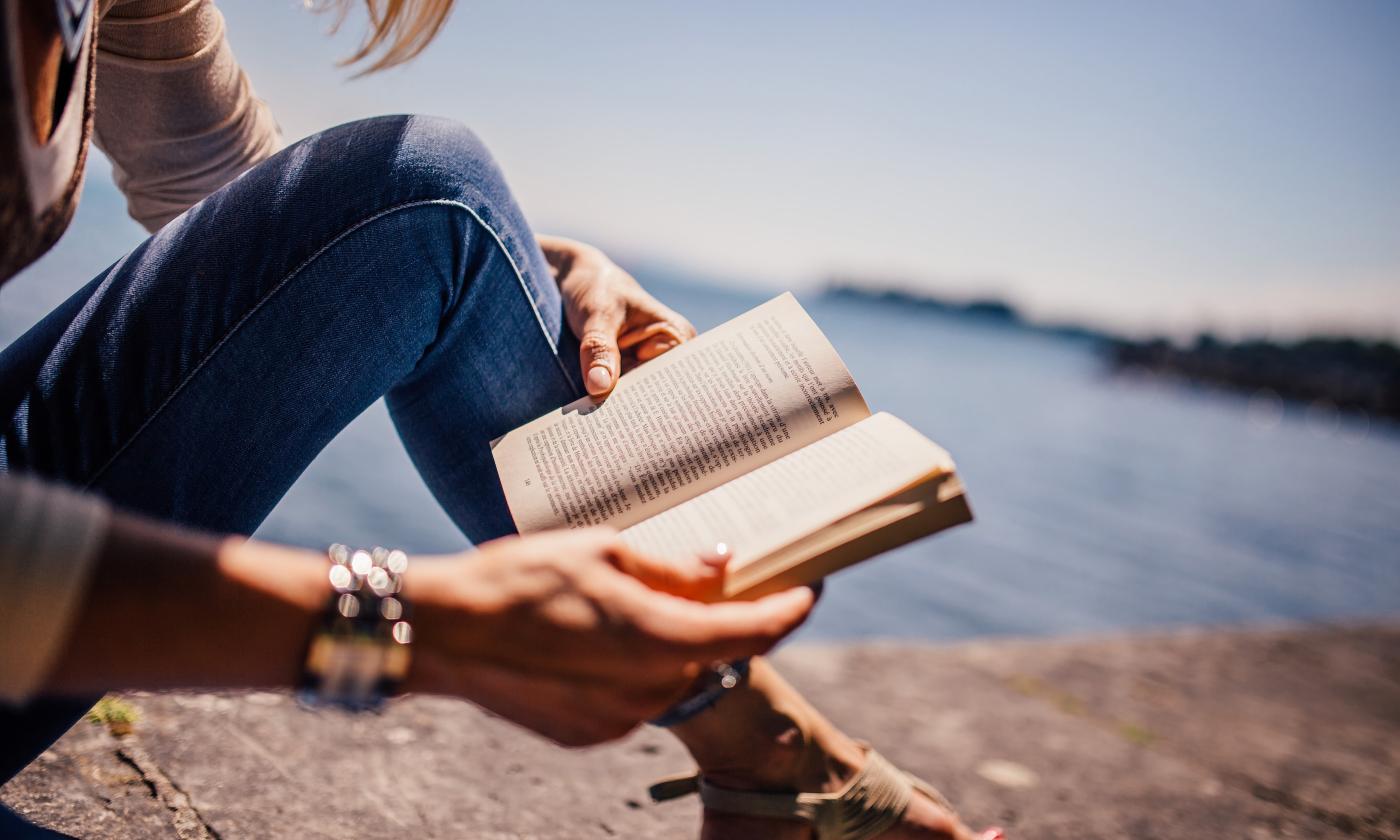 Photo showing woman sign on a stone wall, reading a book with water in the background