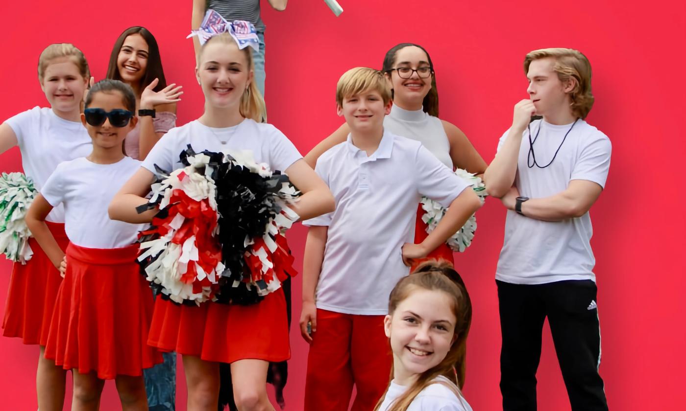 "Miss Beth" cast in cheerleading costumes behind a red backdrop. 