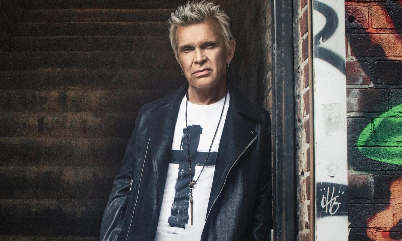 Billy Idol poses in front of a brick backdrop. 