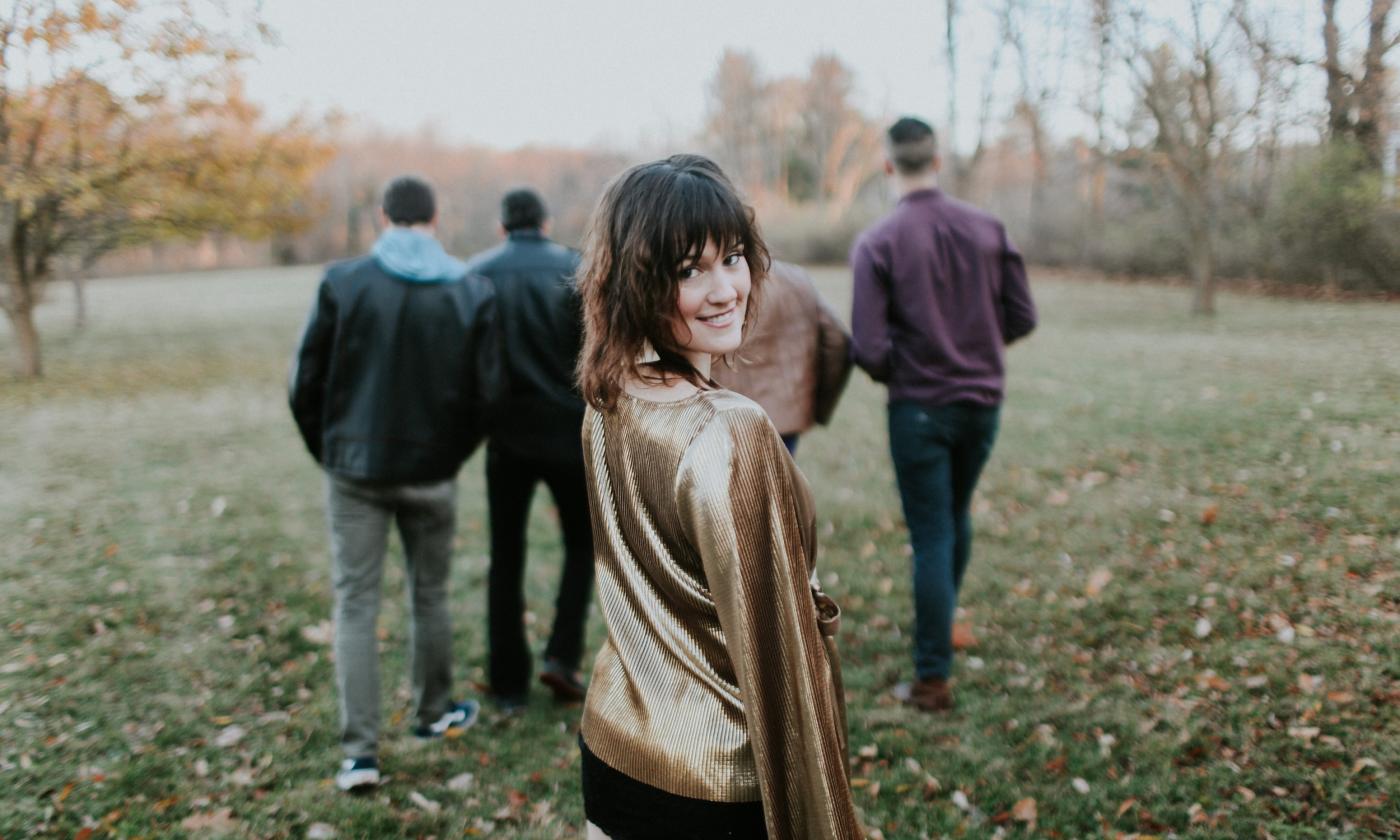 Maddy Walsh looking at the camera while members of The Blind Spots walk in an autumn landscape