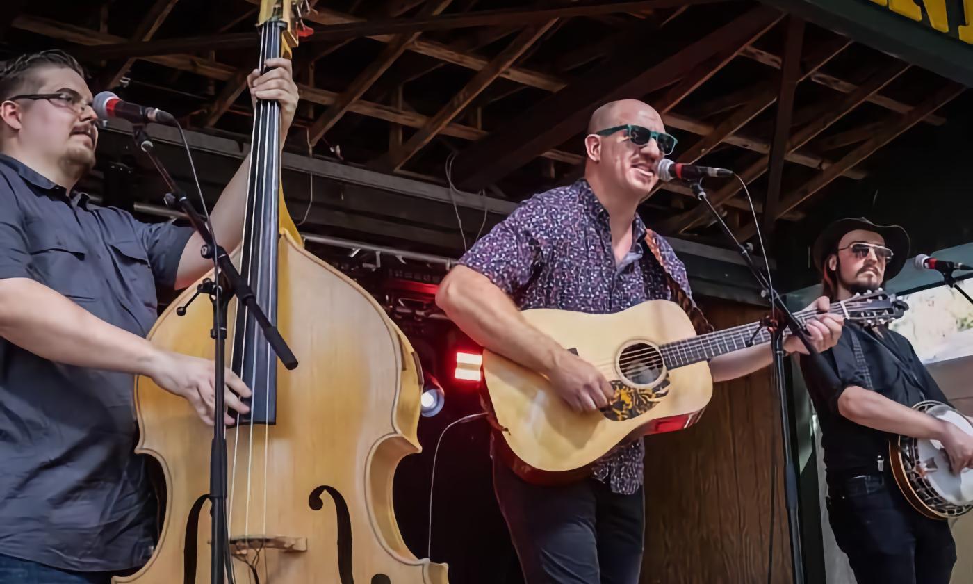 The three members of Grandpa's Cough Medicine, Rex Putnam on bass, Brett Bass on guitar, and Benny McDowell on banjo, performing on stage