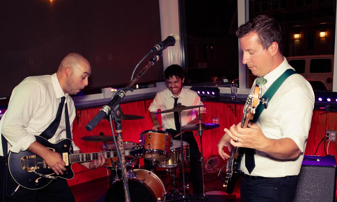 Matt, Andre, and Ian of Ferris Delux performing on stage wearing white shirts, skinny black ties, and black pants