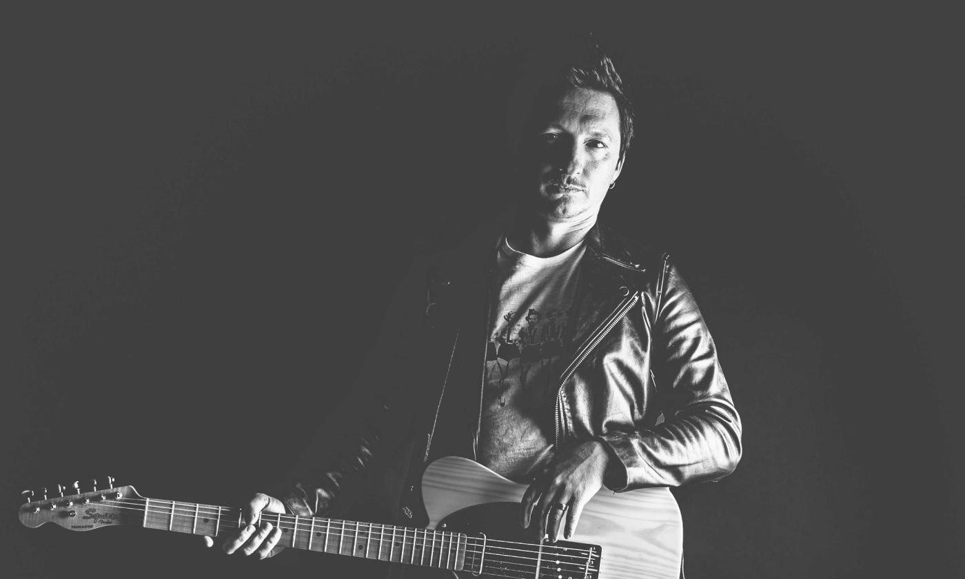Mick Fury, musician, with guitar, and black background behind.