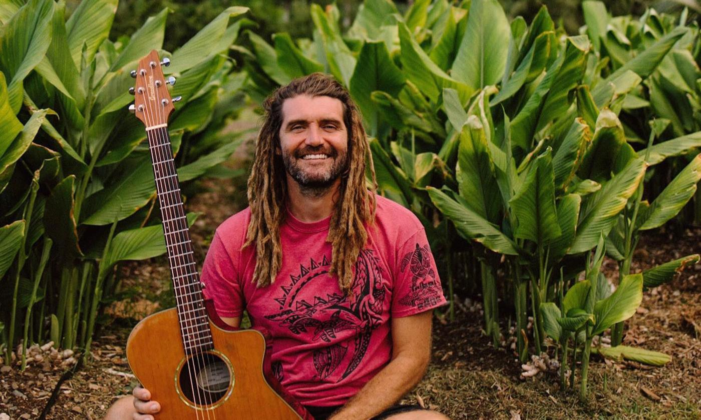 Dinger, songwriter, and conservationist Paul Izak sitting in his garden with a guitar and a big grin on his face. 