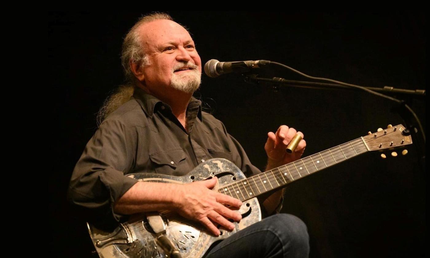 Tinsley Ellis smiles and clutches a guitar in his hand. 