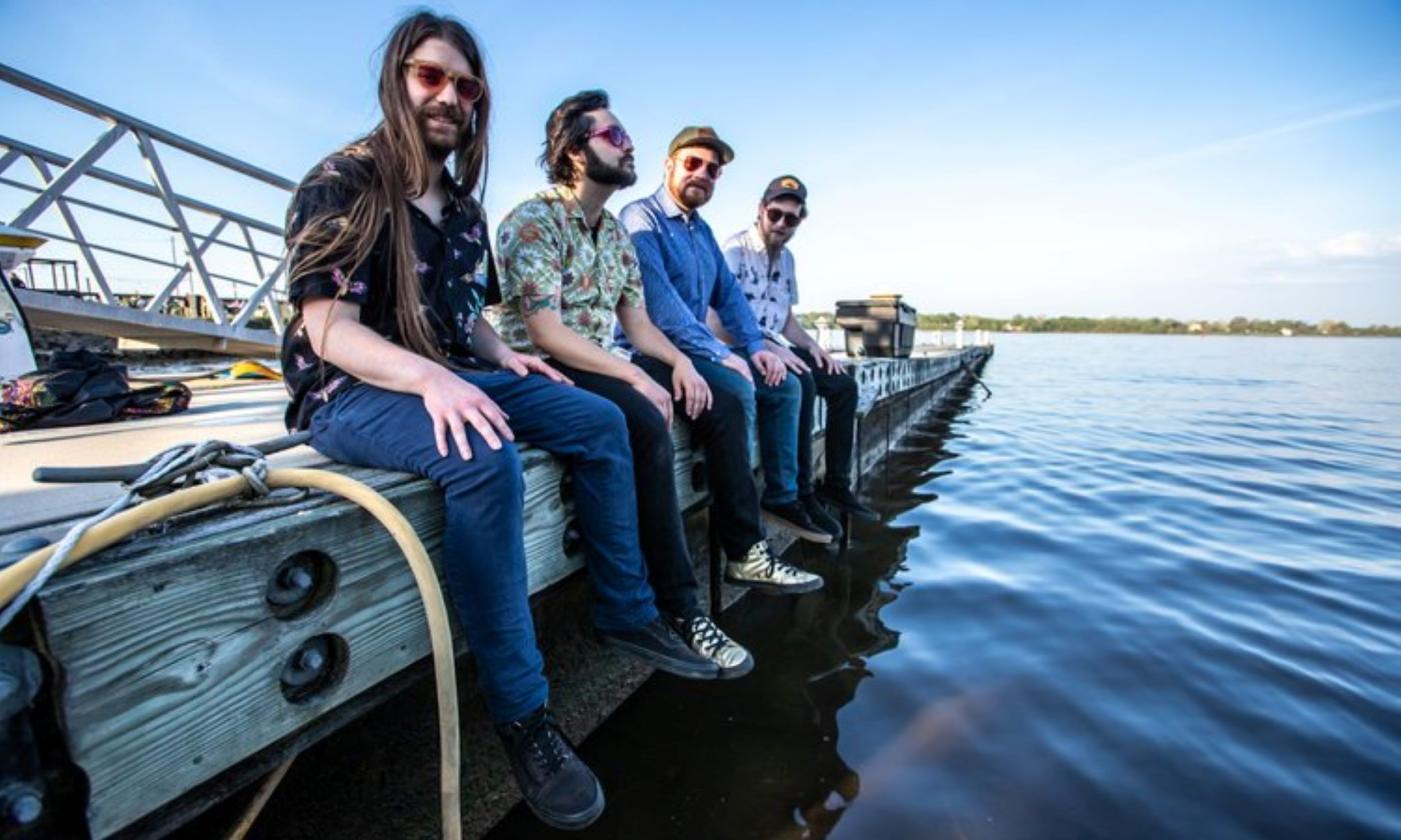 The Dirty Grass Players smile and pose on a dock while placing their feet close to water. 