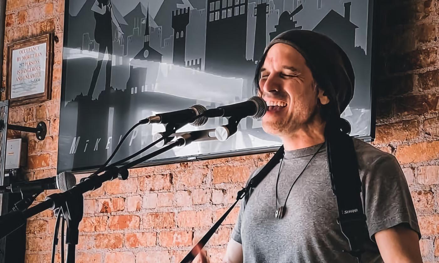 Mikel Paris singing at a microphone in front of a brick wall