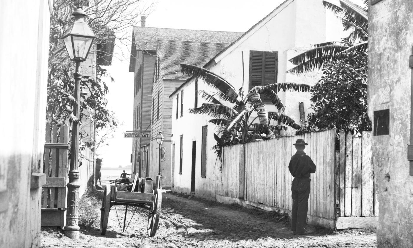 B&W view down Cadiz Street in St. Augustine, circa 1895. Fences lined with banana trees and large colonial houses lead to a view of Matanzas Bay. A young boy faces away from the camera in the foreground.