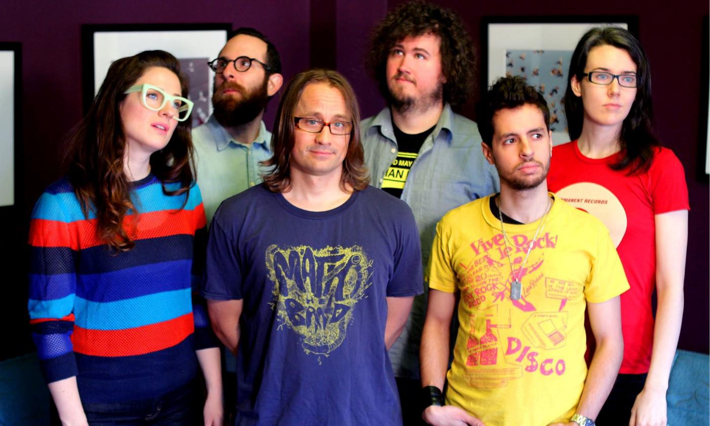Bandmates from Wheatus pose in colorful shirts in front of a wall with framed pictures. 