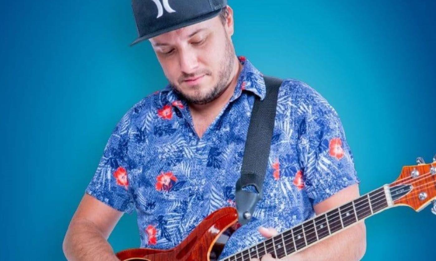 Adam Grayford gazes at his guitar while standing in front of a blue backdrop. 