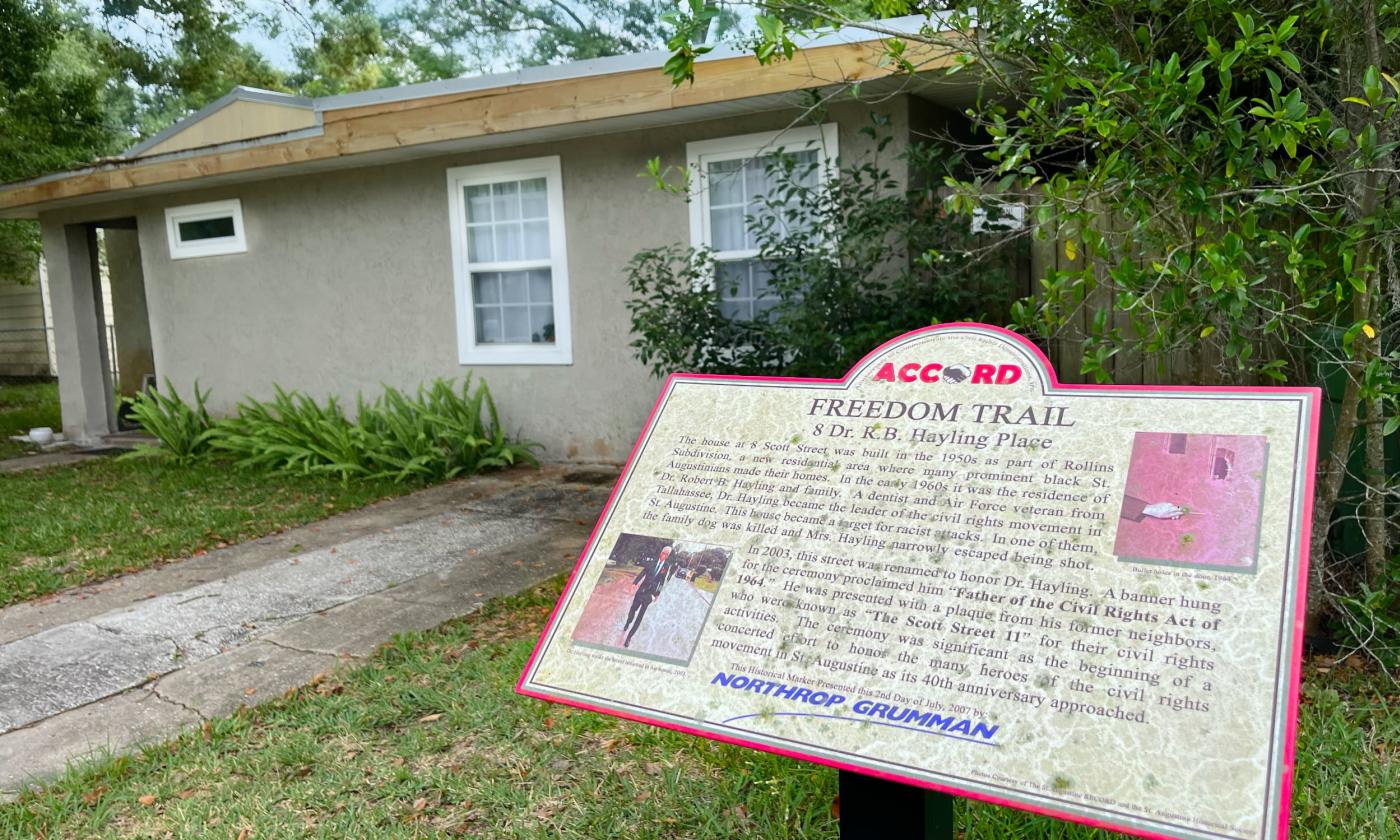 Brown one-story cement block home surrounded by greenery. ACCORD Freedom Trail marker in the foreground.