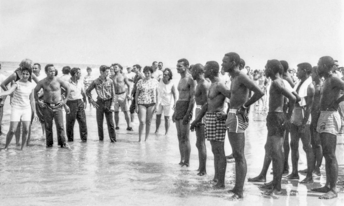 A multi-racial group of people in swimwear, standing in the water at a beach in the 1960s