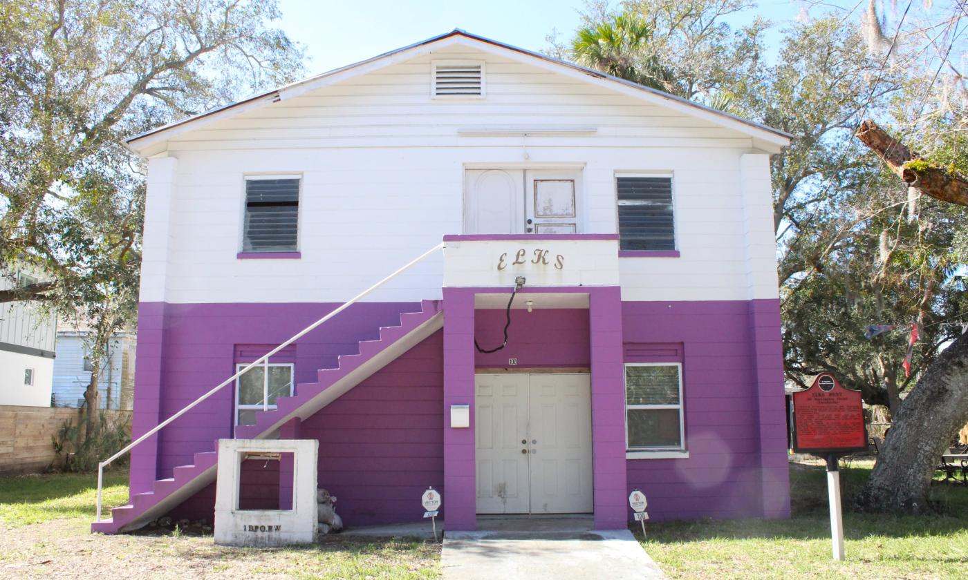 A two story cement block building. Ground floor is painted bright purple, second story painted white. "ELKS" in all caps above the entrance.