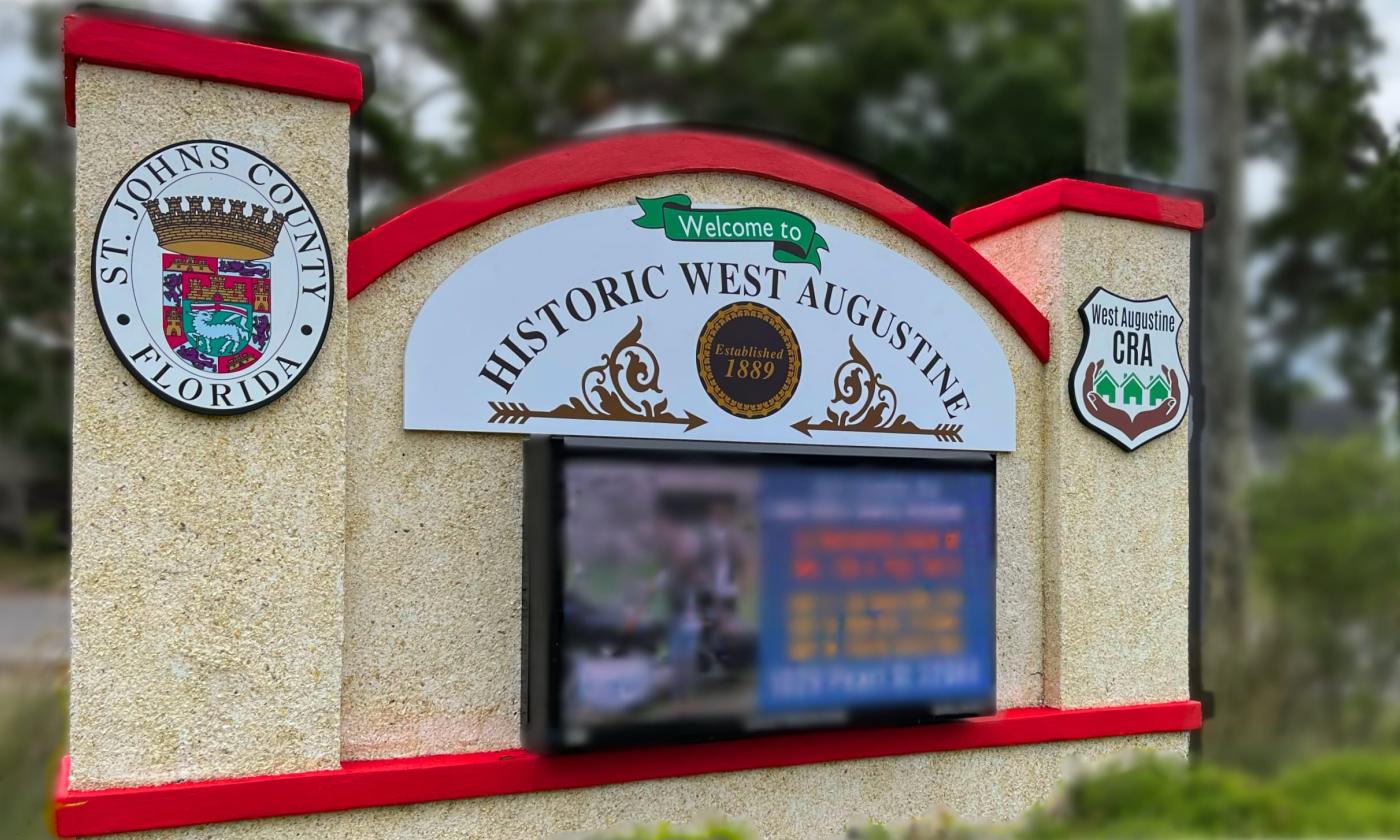 Coquina sign with red details, blurred background of trees. The sign bears the City of St. Augustine emblem, West Augustine CRA emblem, and the words "Historic West Augustine."