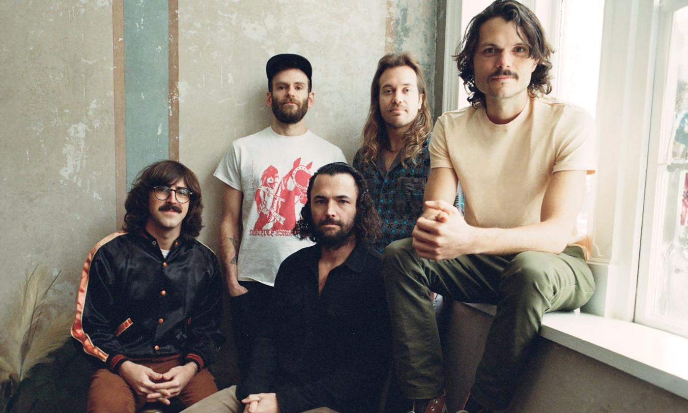 Members of the band Goose pose together near a wall with tan colored wallpaper and a window with a white border. 